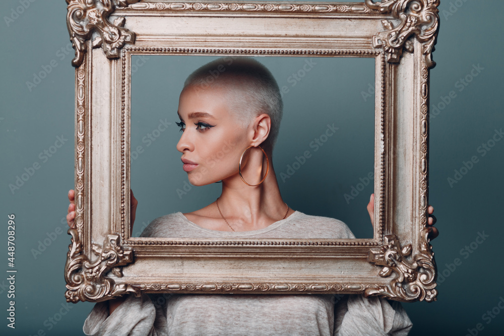 Wall mural millenial young woman with short blonde hair holds gilded picture frame in hands behind her face pro