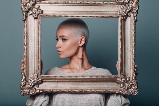 Millenial young woman with short blonde hair holds gilded picture frame in hands behind her face profile portrait