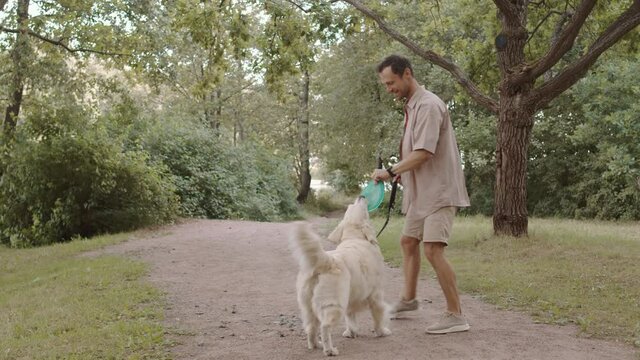 Wide Shot Of Caucasian Man Wearing Casual Outfit, Playing Frisbee With Excited Golden Retriever Dog On Leash In Park On Summer Day