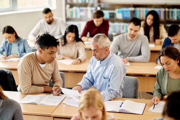 Mature teacher talking to college student while assists him with lecture in classroom.