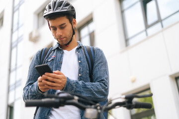 Low-angle view of handsome male courier with backpack standing with bicycle in city street and using navigation app on phone, close-up. Delivery man looking for client address looking smartphone. - obrazy, fototapety, plakaty