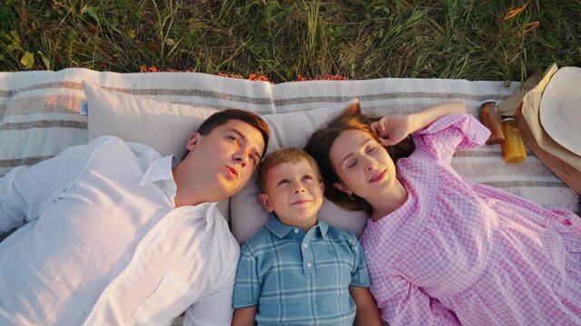 Mother, father and little son lying on blanket, looking up in the sky and telling dreams. Top down family spending time together in nature, rays of sunset on their faces. Concept of bonding
