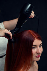 Fototapeta premium Hairdresser working with red-haired model with hairdryer on gray background