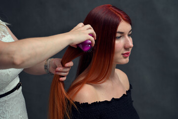 Hands of a hairdresser are working with a girl with red hair on a gray background