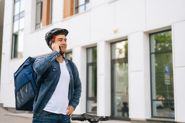 Cheerful Caucasian courier male with thermal backpack talking on mobile phone to customer, looking for address to deliver online order. Deliveryman in protective helmet using smartphone outdoors.