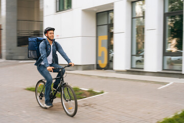 Cheerful handsome young delivery man with thermo backpack riding bicycle in city street on blurred background of office building. Courier male wearing protective helmet delivery food to client