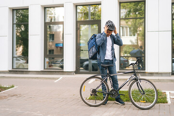 Cheerful courier male getting out of client apartment, wearing helmet, sitting on bicycle and riding off to next delivery. Happy delivery man with large thermo backpack delivery food summer day.