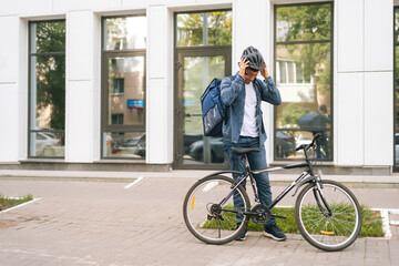 Fototapeta premium Cheerful courier male getting out of client apartment, wearing helmet, sitting on bicycle and riding off to next delivery. Happy delivery man with large thermo backpack delivery food summer day.
