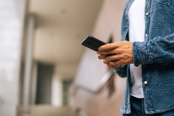 Close-up hands of unrecognizable man texting message on mobile phone outdoors on blurred background, side view, selective focus. Closeup view of male typing sms on modern smartphone outdoors.