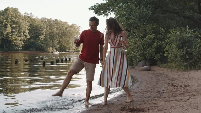 Wide shot of loving Caucasian woman and man holding hands, walking by pond shore, drinking white wine, chatting and smiling