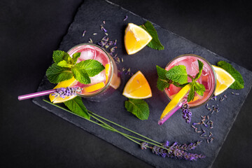 Top view of a fresh summer drink made of lavender, lemon and mint. Refreshing seasonal beverage, served with ice, on black slate board.
