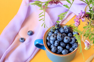 The concept of collecting vitamin berries.Blueberries in a blue cup, light scarf, bokeh from wild flowers, top view, close-up