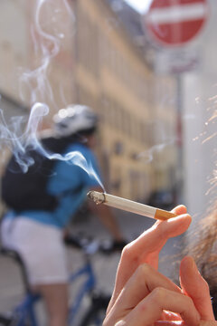Eine Hand hält eine rauchende Zigarette und im Hintergrund ein Fahrradfahrer unscharf