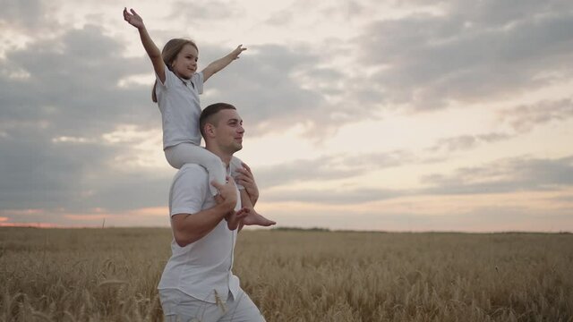 Daddy carries on his shoulders his beloved little healthy daughter in sun. In slow motion, the daughter walks with her father on the field and free and happy waves her hands up. walking in field.