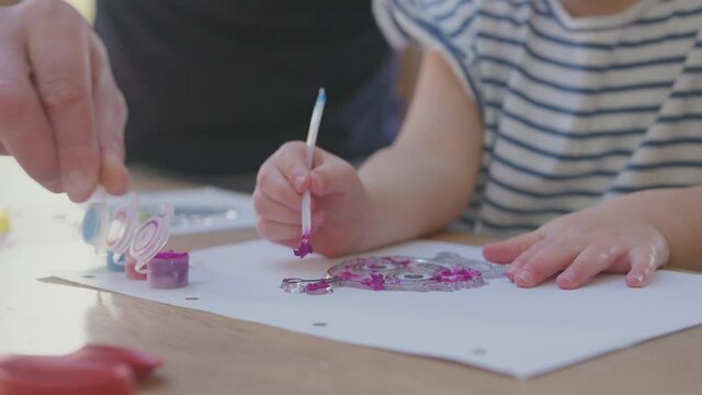 Close up of father and young daughter having fun at home sitting at table and painting decoration together - shot in slow motion