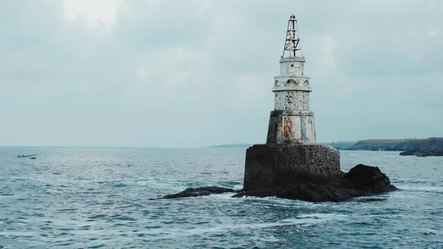 Sea waves hitting and splashing against old white lighthouse and dark gray cliffs. Isolated on blue cloudy sky. Fishing boat passing. Near town of Ahtopol on Black sea coast, Bulgaria