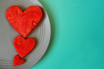 three hearts of different sizes cut from a red urgent watermelon lie on a gray plate close up against a green background. view from above