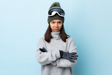 Mixed race skier girl with snowboarding glasses over isolated blue background feeling upset.