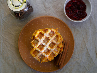 close gluten-free pumpkin Belgian waffles, sprinkled with powdered sugar, lie with cinnamon sticks on a brown plate on a beige table next to currant jam top view