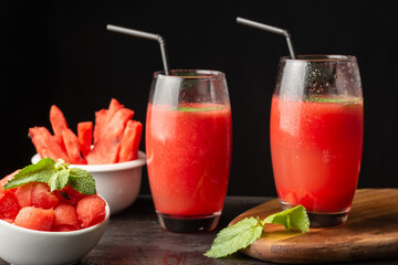 View of two glasses with watermelon juice, straw and bowls with pieces of watermelon, selective focus, on wooden table and black background, horizontal