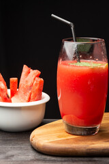 Close-up of glass with watermelon juice with mint and straw, bowl with pieces of watermelon, on wooden table, black background, vertical