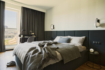 Stylish composition of small modern bedroom interior. Bed, creative lamp and elegant personal accessories. Walls with black panels. Panoramic windows. Minimalistic masculine concept. Template.