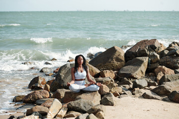 Calm young sportswoman meditating on rocky beach, she is sitting in lotus position with eyes closed