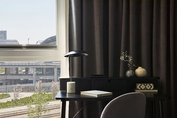 Stylish composition of small modern workspace interior with lamp and personal accessories. Walls with black panels. Panoramic windows with grey curtains. Minimalistic masculine concept. Template.