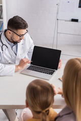 Pediatrician looking at laptop with blank screen near blurred child and parent in hospital