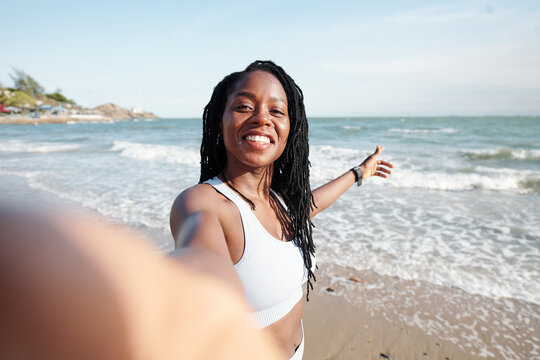 Pretty young woman with dreadlocks smiling and taking selfie on beach