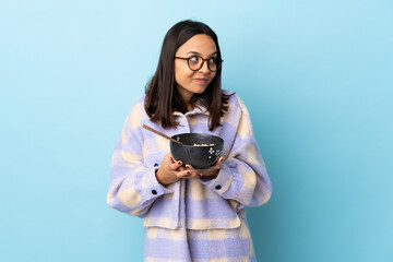 Young brunette mixed race woman holding a bowl full of noodles over isolated blue background...