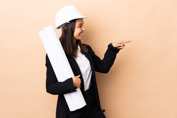 Young architect woman holding blueprints over isolated background pointing finger to the side