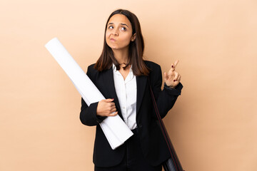 Young architect woman holding blueprints over isolated background with fingers crossing and wishing the best