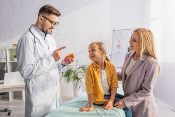 Obraz premium Smiling doctor holding pills and pointing with finger at mother and kid on medical couch