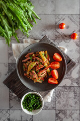Fried potatoes with ham in a flat gray plate with cherry tomatoes and green onions on a tiled gray background, top view