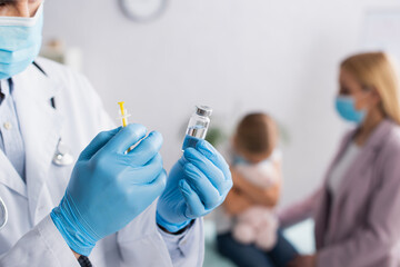 Doctor in medical mask holding vaccine near blurred mother and kid