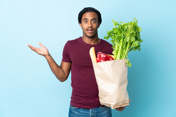 African American man holding a grocery shopping bag isolated on blue background with shocked facial...