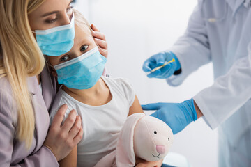 Mother hugging kid in medical mask near pediatrician with syringe on blurred background