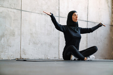 Young muslim woman in hijab meditating while sitting on fitness mat indoors