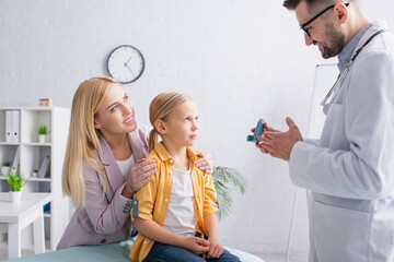 Smiling mother hugging child near family doctor with inhaler in clinic