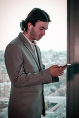 The young trader looks at his cell phone next to a window. Behind him, a large city can be seen.
