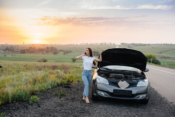 A young girl stands near a broken-down car in the middle of the highway during sunset and tries to call for help on the phone. Breakdown and repair of the car. Waiting for help.