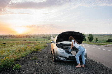 A young girl stands near a broken car in the middle of the highway during sunset and tries to repair it. Breakdown and repair of the car. Troubleshooting the problem.