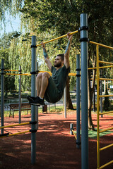 Active lifestyle concept. Young handsome Caucasian man pulls himself up on street sports field on warm summer morning. Strength training for muscles of arms. Hang on horizontal bar and lift legs.