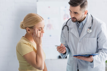 doctor with clipboard talking to woman touching head with closed eyes