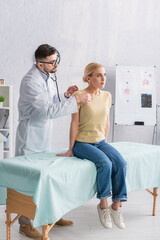 woman in jeans sitting on medical couch while doctor examining her with stethoscope