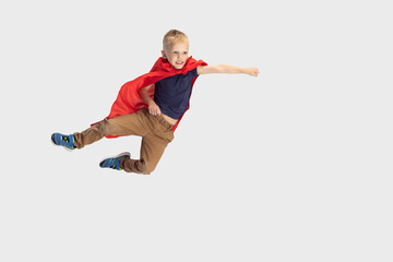 Portrait of little blond boy, pupil flying like super hero in red cape isolated on white studio background. Copyspace for ad. Childhood, education, emotion concept.