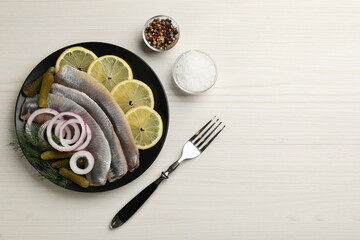 Salted herring fillets served with onion rings, pickles, dill and lemon on light beige wooden table, flat lay. Space for text