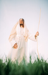 Jesus Christ with wooden staff stands clothed in white robe against sky.