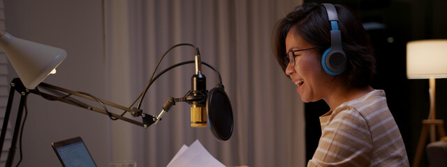 Asian female podcaster recording and broadcasting her podcast from home. Woman working from home at night reading a notepad, paper work and talking into a microphone.